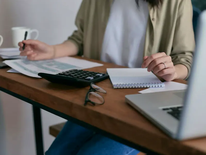 A woman manages finances at home, using a laptop and calculator on a wooden desk. | Immagine di Mikhail Nilov, su https://www.pexels.com/@mikhail-nilov?utm_source=centralapp&utm_medium=referral