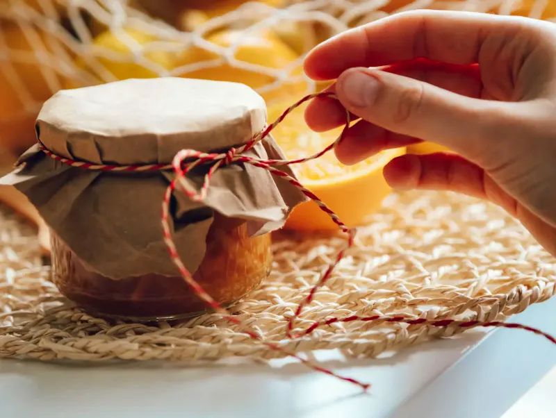 Homemade jam jar with hand tying string, surrounded by fresh oranges in warm lighting. | Immagine di ROMAN ODINTSOV, su https://www.pexels.com/@roman-odintsov?utm_source=centralapp&utm_medium=referral