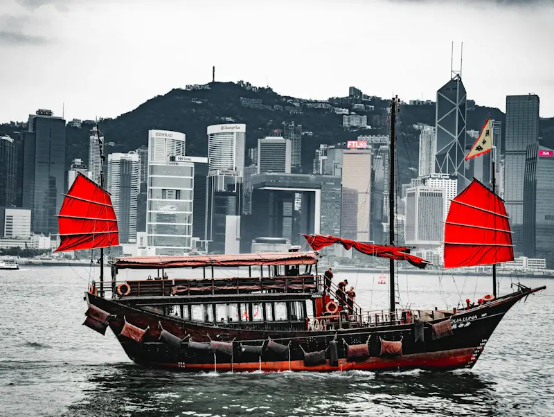 A traditional junk boat with red sails in Hong Kong harbor, against a skyline backdrop. | Photo by Nitin Sharma, on https://www.pexels.com/@tasveerwala?utm_source=centralapp&utm_medium=referral