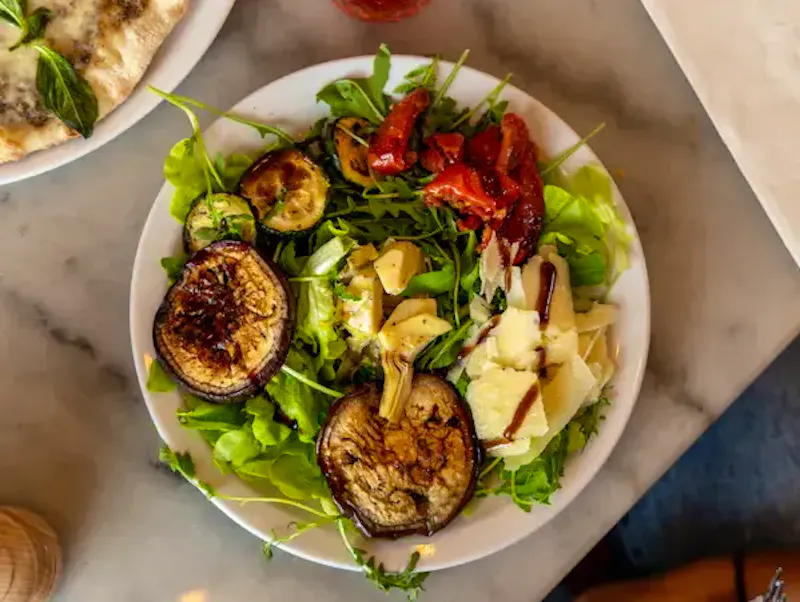Une entrée italienne colorée composée de légumes du soleil (aubergines, courgettes, artichauts) et de tomates confites sur un lit de salade, le tout parsemé de copeaux de parmesan frais chez Ti Amo.